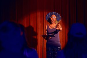 A Black woman with a wide hat performs a spoken word on a stage illuminated in red light