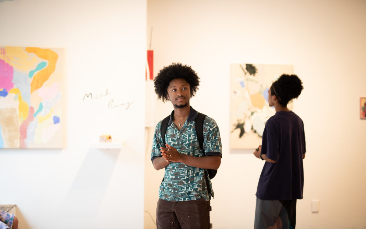 An African American man stands in the center of this photo. He is wearing a blue patterned shirt and a backpack. He is in a bright gallery and few paintings are visible in the background.