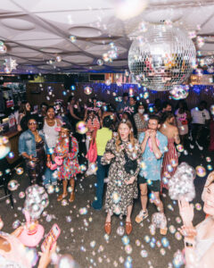 a crowd dances under a disco ball as bubbles float around