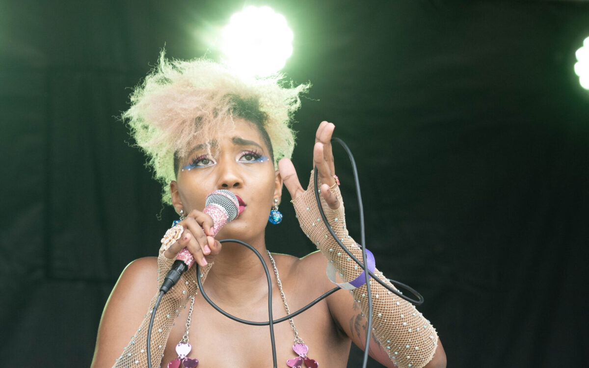 A black female performer with short blonde hair sings into a microphone. She is wearing blue flare pants