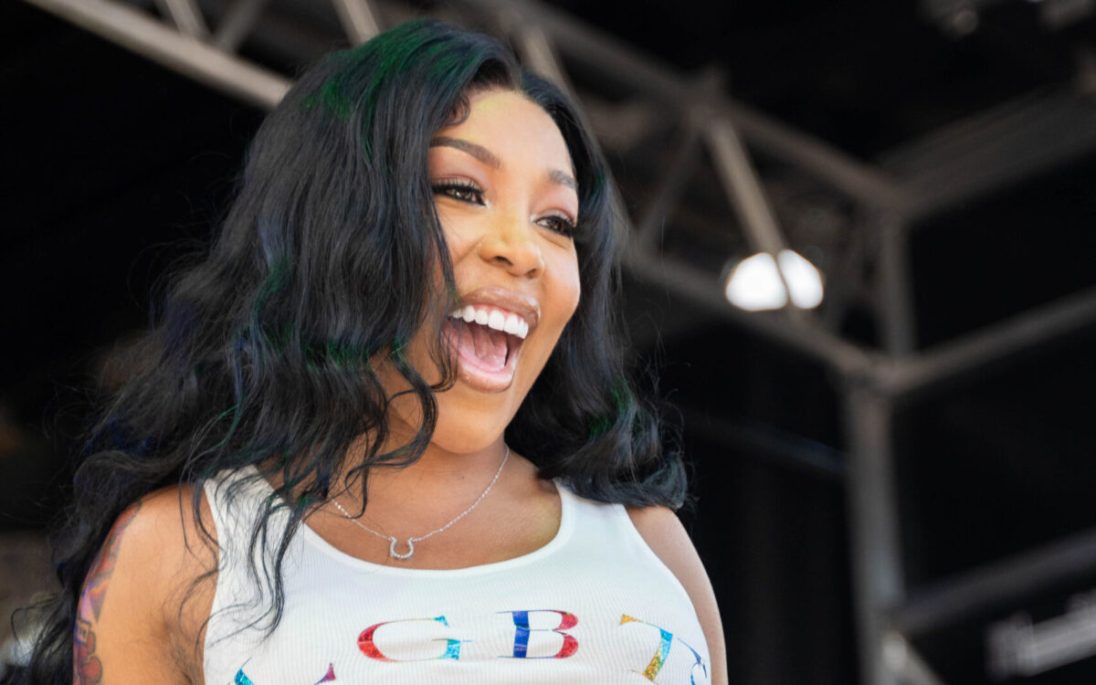 A Black woman stands on stage in a white tank top and jean shorts